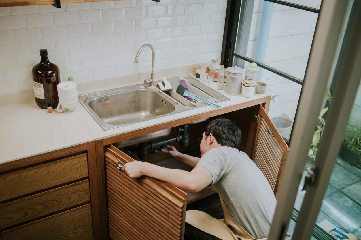 A person performing plumbing maintenance under a kitchen sink, inspecting pipes, and ensuring everything is in working order.
