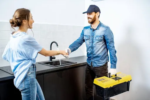 Homeowner shaking hands with a plumber carrying a toolbox in a kitchen.