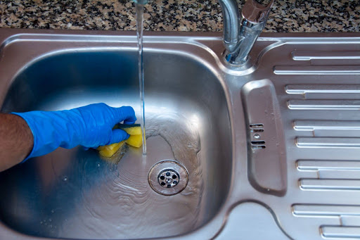 Zoomed in shot of someone cleaning a stainless steel kitchen sink with an orange and green sponge and wearing a blue cleaning glove.