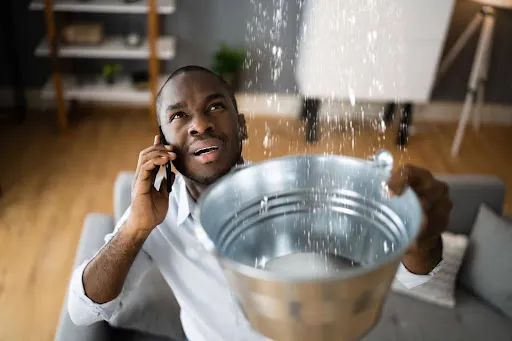 Homeowner calling for help while holding a metal bucket beneath water pouring from the ceiling.