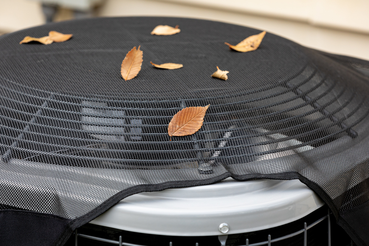 Close-up of an outdoor air conditioner condenser with a protective mesh cover and leaves on top.